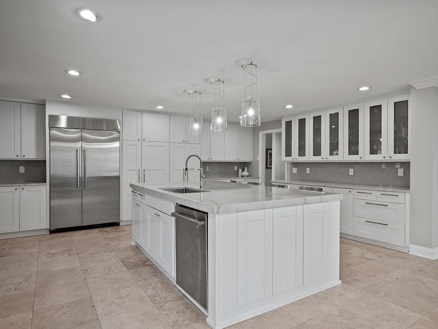 This is a bright, modern kitchen featuring white cabinetry, stainless steel appliances, and a large central island with a light-colored countertop. The kitchen is well-lit with recessed lighting and pendant lights above the island, creating a clean and inviting atmosphere. The perspective is from a wide angle, showcasing the entire kitchen space.