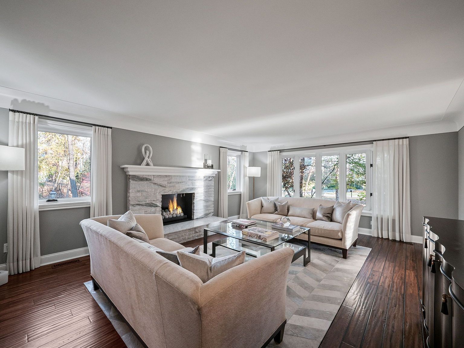 This is a well-lit living room featuring two beige sofas, a glass coffee table, and a fireplace with a marble surround. Large windows with white curtains provide ample natural light, and the dark hardwood floors add warmth to the space. The room is decorated in a neutral color palette, creating a comfortable and inviting atmosphere.