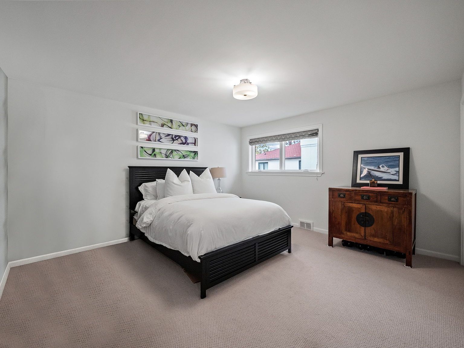 This is a well-lit bedroom featuring a dark wood bed with white linens and pillows. Above the bed are three framed pieces of art. To the right of the bed is a wooden dresser with a framed picture on top. The room has neutral-colored carpet and white walls, creating a clean and inviting space.