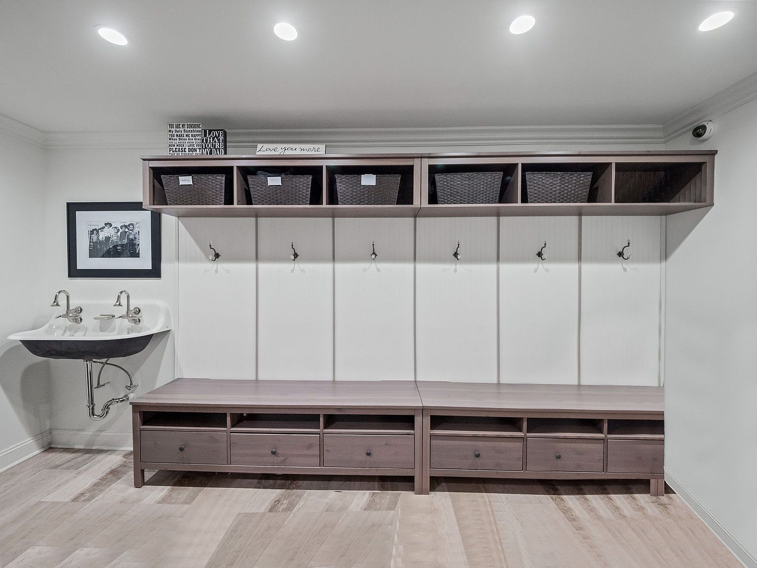 This interior shot showcases a well-organized laundry room or mudroom area. It features a utility sink with vintage-style faucets, a built-in bench with drawers, and a storage unit with baskets above coat hooks. The room is decorated with a framed picture and decorative signs, creating a functional yet stylish space.