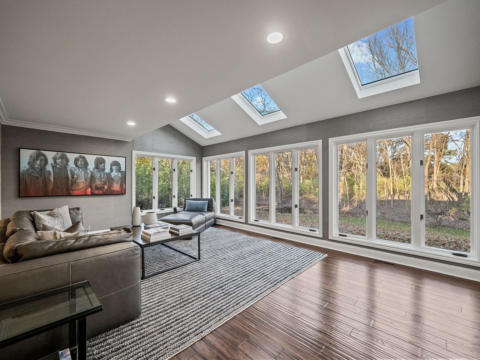 This is a bright and airy living room featuring a modern design with a gray color scheme. The room is well-lit with natural light from multiple windows and skylights, showcasing the outdoor scenery. A comfortable leather sofa, a stylish coffee table, and a large piece of art add to the room's appeal, while a striped rug anchors the space.
