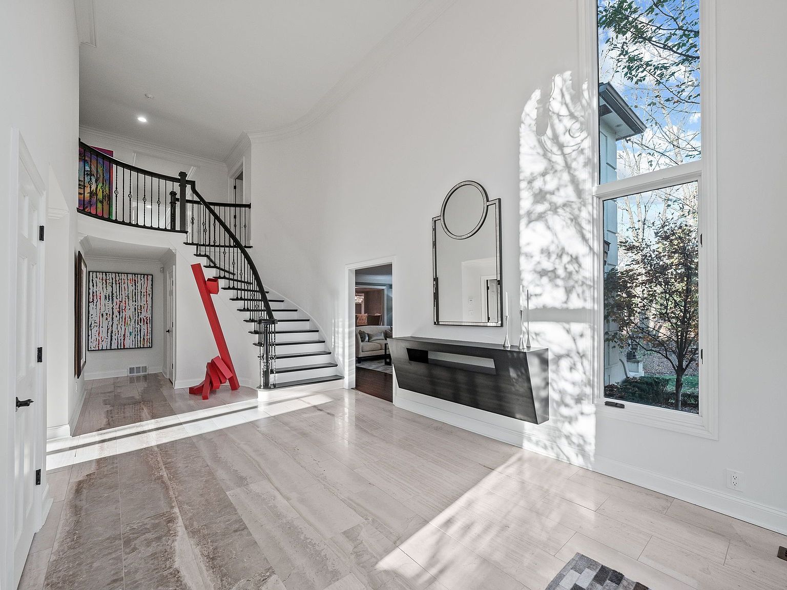 This is a grand foyer featuring a curved staircase with black iron railings and white risers. A large window provides ample natural light, highlighting the light-colored hardwood floors. A modern console table and mirror add a touch of elegance to the space, while a red sculpture adds a pop of color.