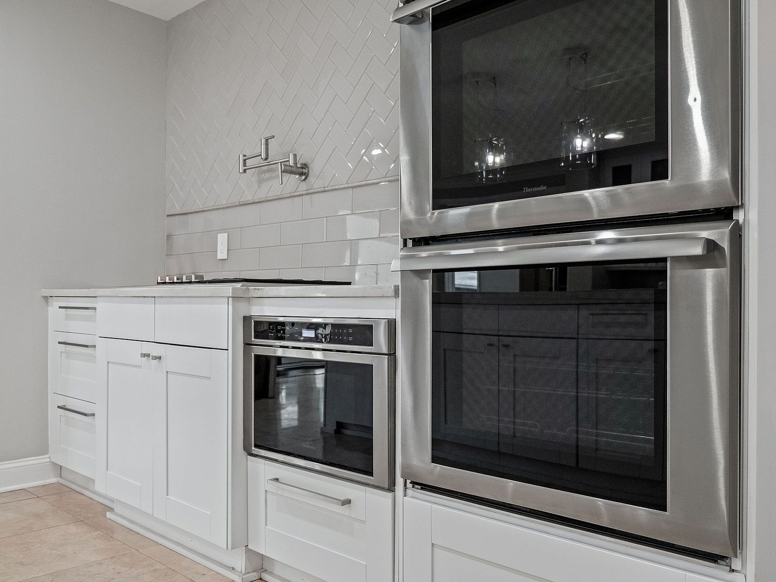 This is a well-lit kitchen featuring white cabinets and stainless steel appliances, including a double oven and a built-in microwave. The backsplash is a light gray tile in a herringbone pattern, adding a touch of modern elegance. The perspective is a medium shot, focusing on the appliances and cabinetry.