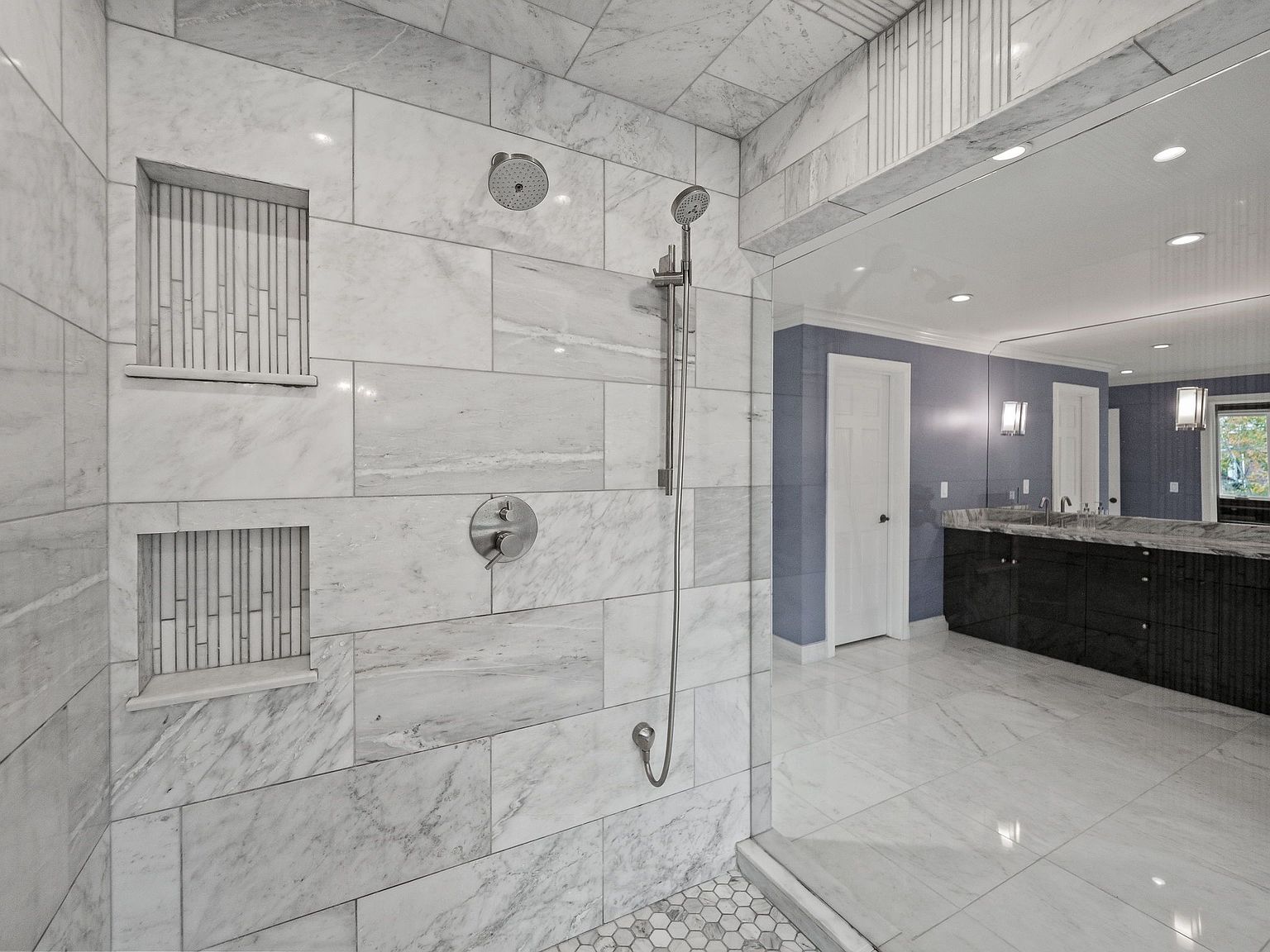 This is a luxurious primary bathroom featuring a walk-in shower with marble tiling, dual shower heads, and built-in niches. The bathroom also includes a vanity with dark cabinetry, a light countertop, and a large mirror. The overall impression is clean, modern, and high-end.