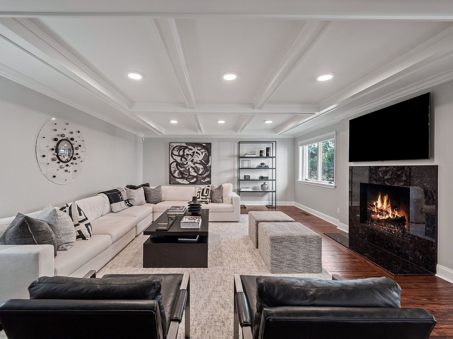 This is a well-lit living room featuring a large sectional sofa with decorative pillows, a dark coffee table, and two black leather chairs in the foreground. A modern fireplace with a mounted TV above it serves as a focal point, complemented by a bookshelf and abstract art on the walls. The room is decorated in neutral tones with hardwood floors and a coffered ceiling, creating a sophisticated and inviting atmosphere.