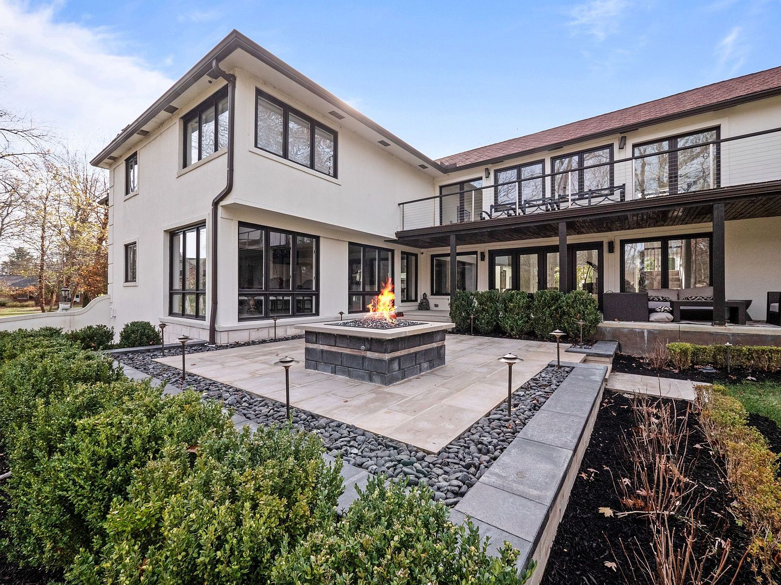 This image showcases the rear exterior of a modern two-story home, featuring a patio with a fire pit and manicured landscaping. The house has large windows with dark frames, a second-story balcony, and a combination of stucco and siding. The outdoor space is designed for relaxation and entertainment, enhancing the property's appeal.