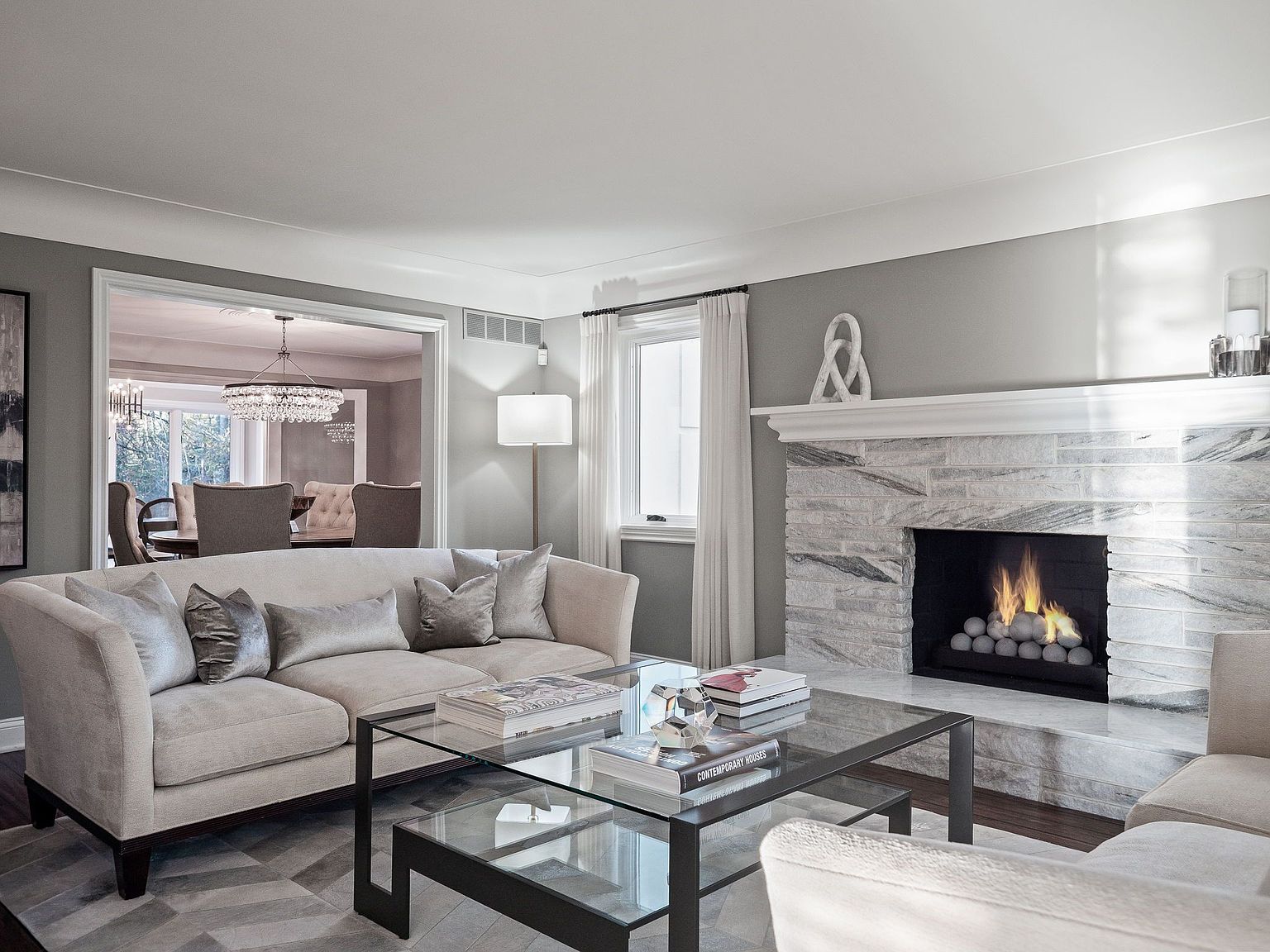 This is an interior shot of a living room featuring a neutral-toned sofa with decorative pillows, a glass-topped coffee table with books, and a fireplace with a stone surround. The room is painted in a soft gray, and a doorway leads to a dining area with a chandelier. The overall impression is elegant and inviting.