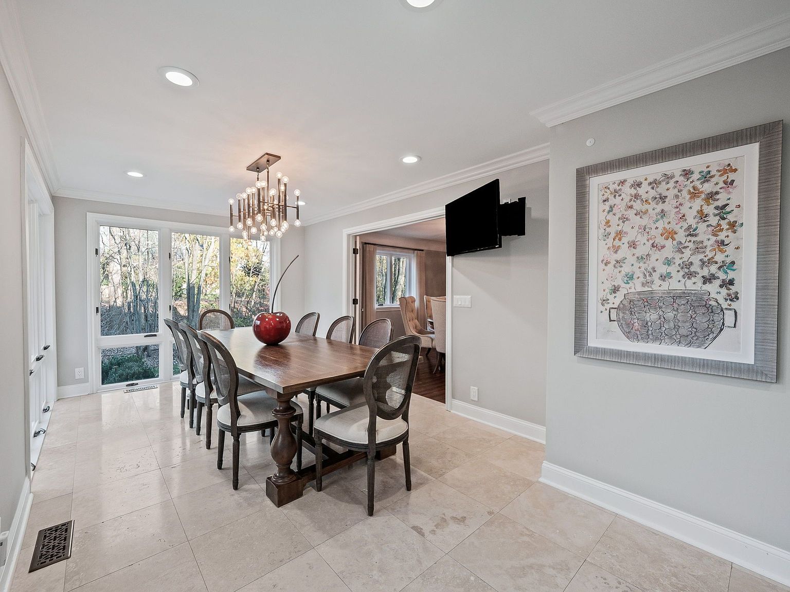 This is an interior shot of a dining room featuring a large wooden dining table with upholstered chairs. A modern chandelier hangs above the table, and a large window provides natural light. A piece of framed art hangs on the wall, and a television is mounted nearby. The room has a neutral color palette and a clean, elegant style.