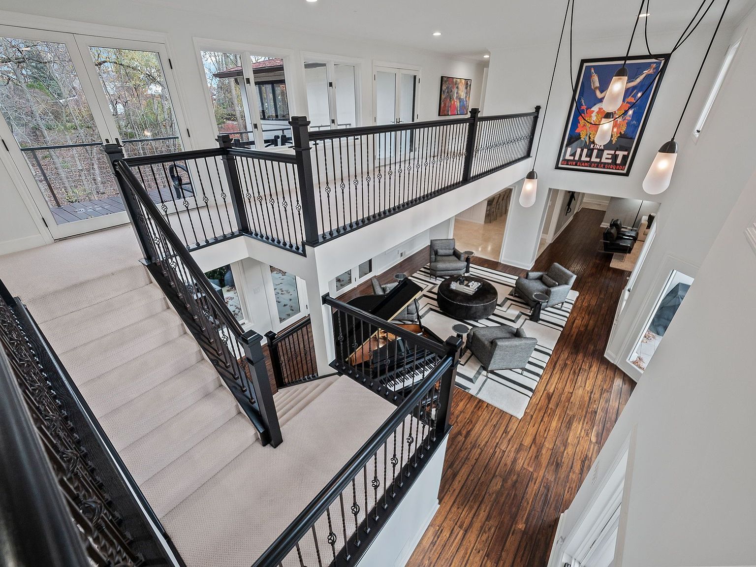 This is an interior shot of a grand foyer featuring a staircase with black wrought iron railings and light-colored carpeting. The view is from above, looking down into the living area with hardwood floors, a striped rug, and a seating arrangement with gray armchairs and a round ottoman. A large poster hangs on the wall, and pendant lights add to the modern aesthetic.