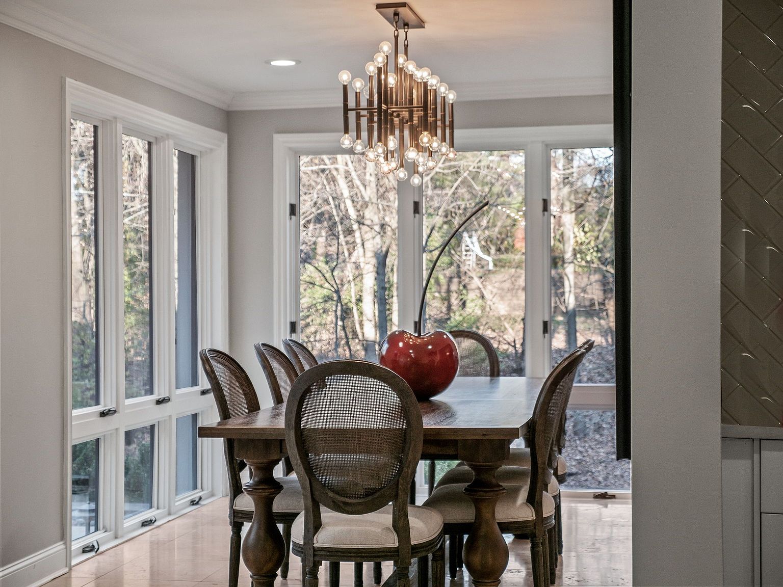 This is an interior shot of a dining room featuring a wooden dining table with six chairs. A modern chandelier hangs above the table, and a large red cherry sculpture sits in the center. Large windows provide natural light and a view of the outdoors, creating a bright and inviting space.