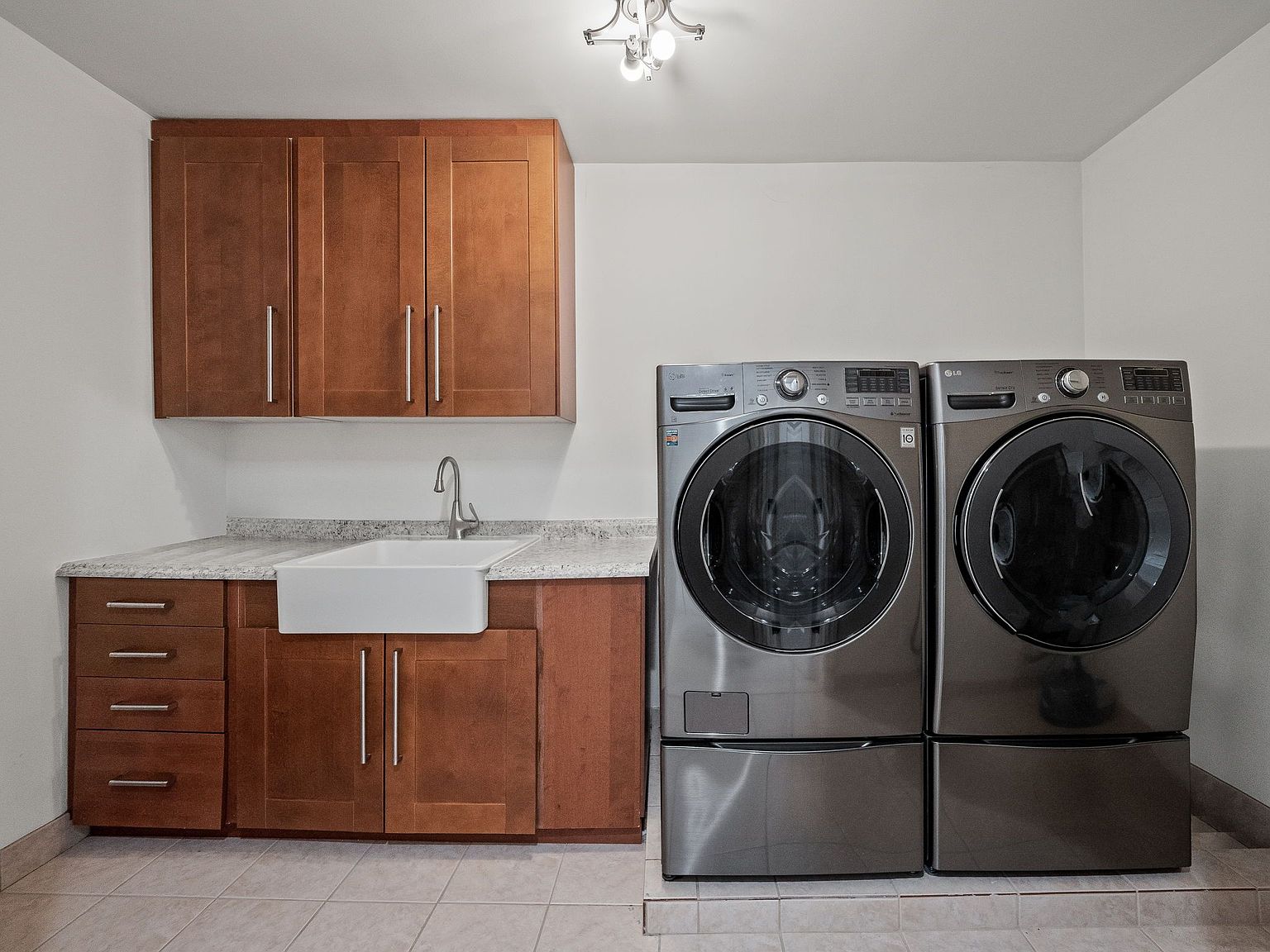 This is a well-organized laundry room featuring a front-loading washer and dryer set in a dark metallic finish, positioned side-by-side. Above the appliances, there are wooden cabinets providing ample storage space, complemented by a sink and countertop area. The room is brightly lit, creating a clean and functional space.