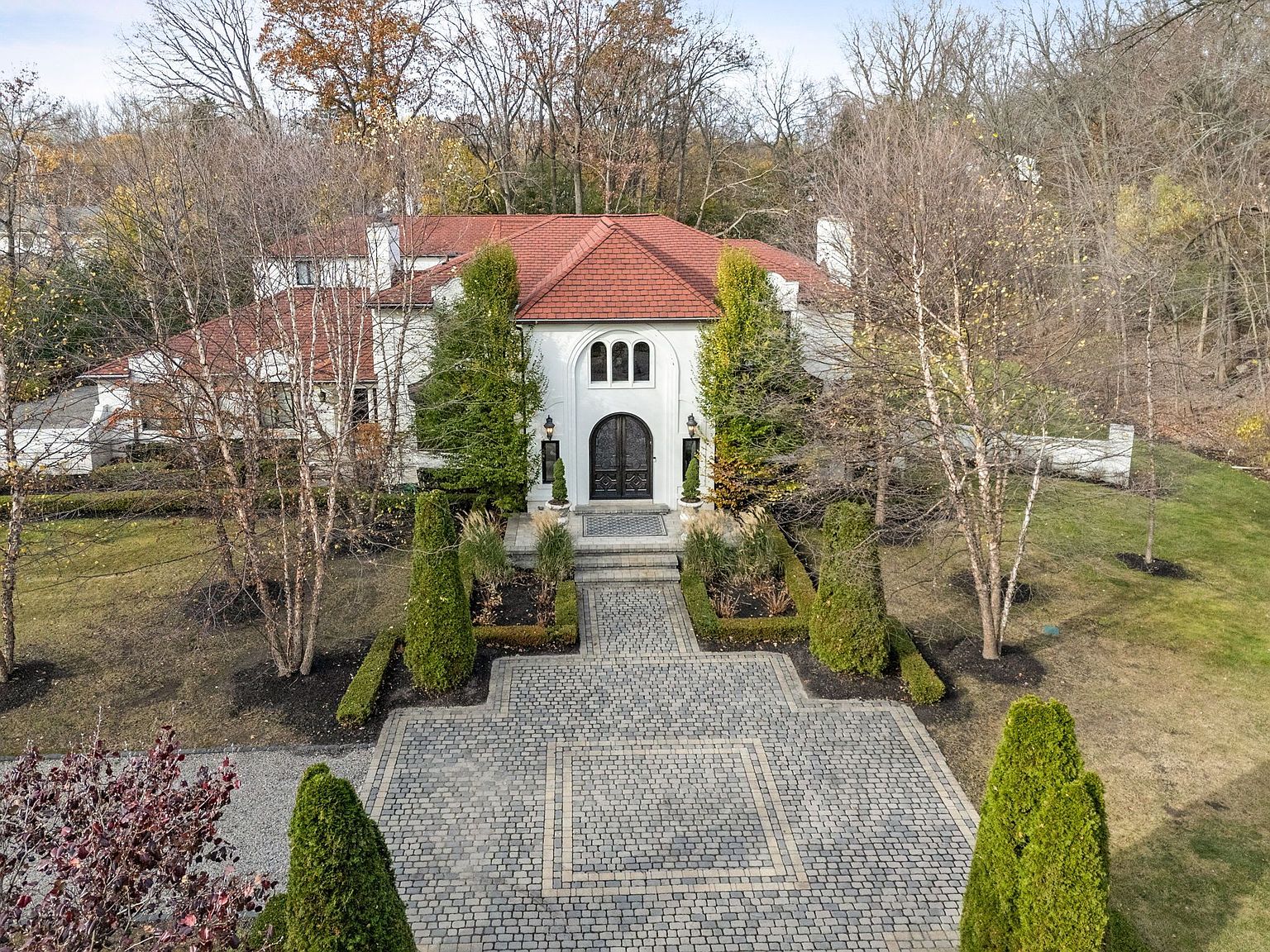 This aerial view showcases a grand, white stucco home with a striking red tile roof, framed by mature trees and manicured landscaping. A patterned cobblestone driveway leads to the arched entryway, flanked by symmetrical plantings and elegant outdoor lighting. The overall impression is one of luxury and sophisticated curb appeal.