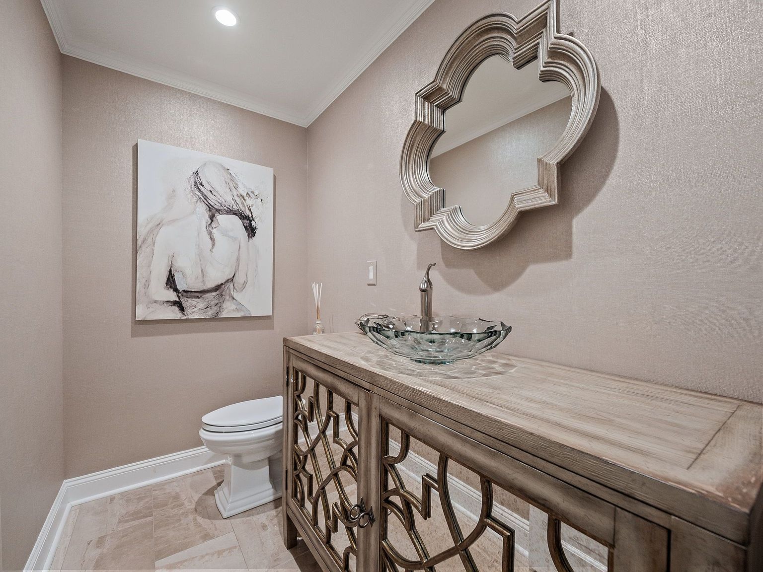 This is a well-decorated guest bathroom featuring a decorative mirror above a vanity with a glass bowl sink and a modern faucet. A piece of art hangs above the toilet, adding a touch of elegance to the space. The color palette is neutral, creating a calming and sophisticated atmosphere.