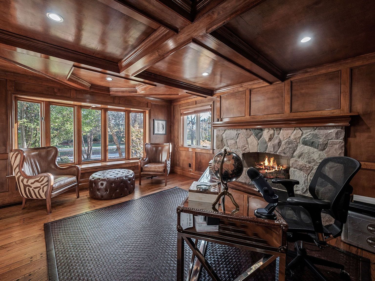This is a luxurious home office featuring rich wood paneling, a coffered ceiling, and a stone fireplace. A large window provides natural light, and the room is furnished with leather chairs, a desk with a globe, and a patterned rug. The overall impression is one of sophistication and comfort, ideal for a productive and relaxing workspace.
