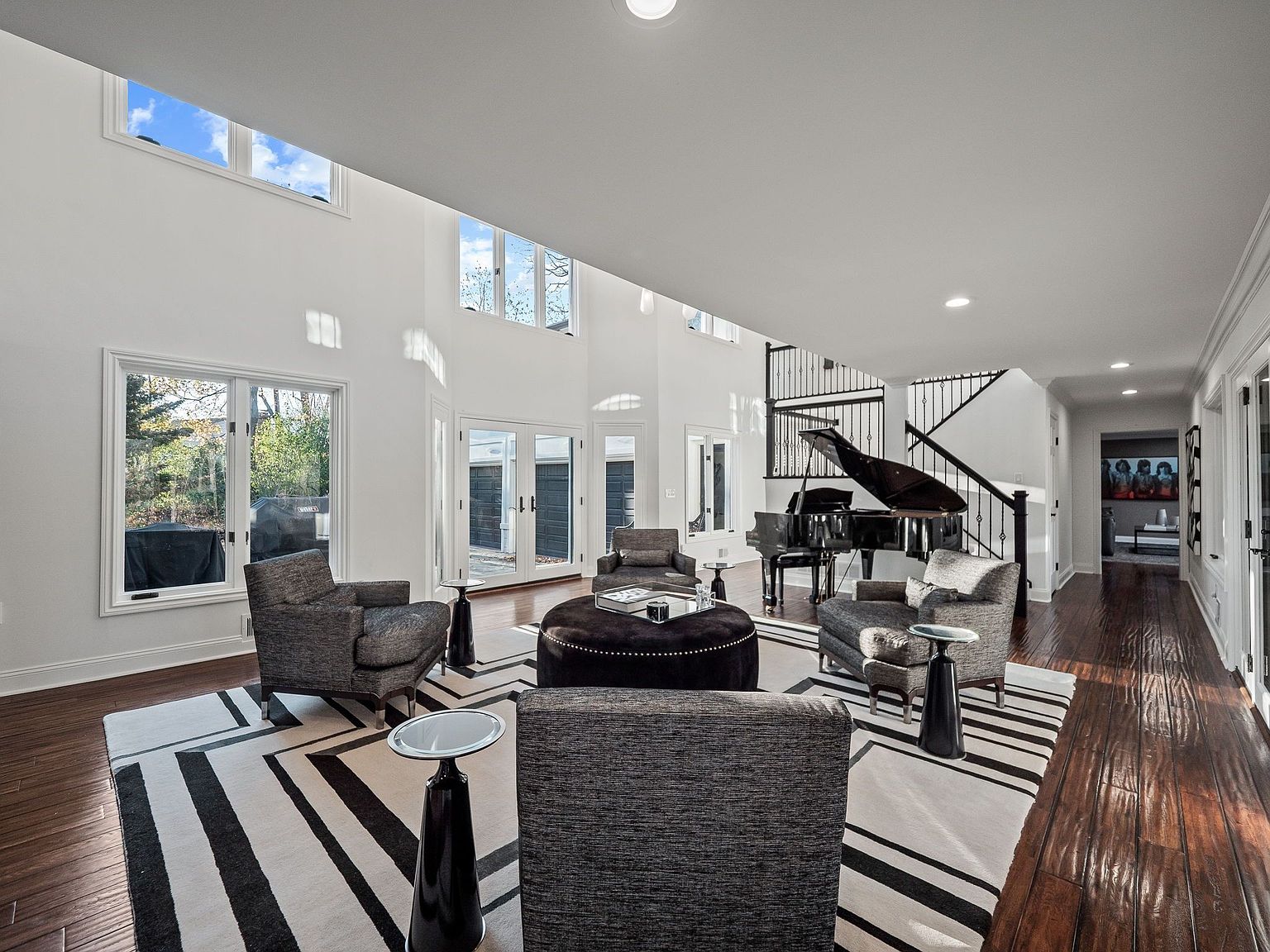 This is a spacious living room featuring high ceilings and an open floor plan. The room is furnished with modern seating arrangements around a central ottoman, complemented by a grand piano near the staircase. A striking black and white striped rug anchors the space, adding a touch of contemporary elegance.