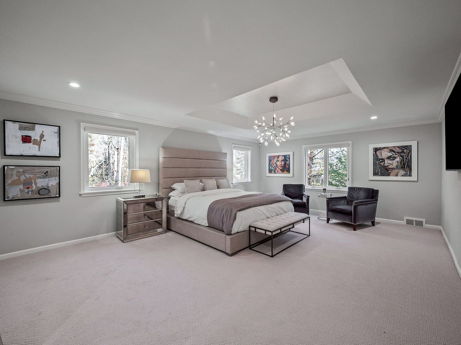 This is a spacious primary bedroom featuring a large upholstered bed with a padded headboard, flanked by mirrored nightstands. The room is decorated with modern artwork and includes a seating area with two armchairs and a bench. The neutral color palette and ample natural light create a serene and luxurious atmosphere.