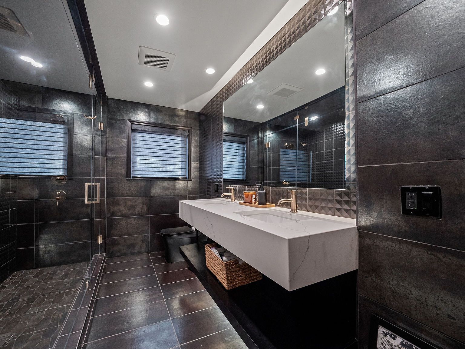 This is a modern primary bathroom featuring dark tile walls and flooring, a large mirror with decorative trim, and a floating double vanity with a white countertop. The bathroom includes a glass-enclosed shower and a window with blinds, creating a sleek and contemporary aesthetic. The perspective is from the doorway, showcasing the entire space.