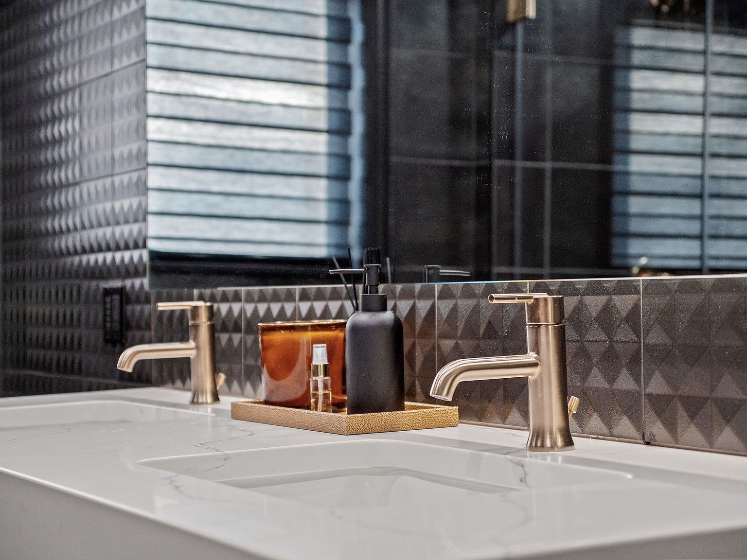 This is a close-up shot of a modern primary bathroom featuring a double vanity with white countertops and brushed gold faucets. A decorative tray holds a candle and toiletries, adding a touch of luxury. The backsplash is dark and textured, reflecting in the mirror behind the vanity.