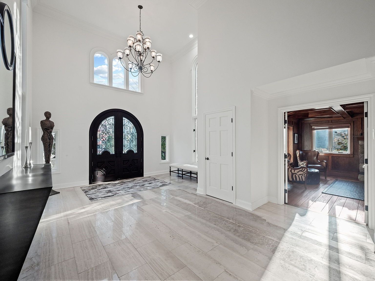 This is an interior shot of a grand foyer featuring a high ceiling, light-colored marble flooring, and a large black arched front door with decorative glass. A chandelier hangs from the ceiling, and there is a glimpse into a wood-paneled room through an open doorway. The perspective is from the entrance, showcasing the spaciousness and elegant design of the home.