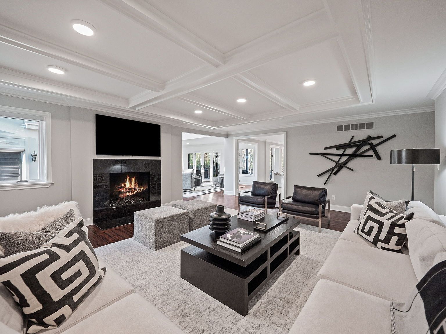 This is a well-lit living room featuring a modern design with a fireplace and a large television mounted above it. The room includes a neutral-colored sofa with decorative pillows, two armchairs, a dark-colored coffee table, and a light-colored area rug. The ceiling has a coffered design with recessed lighting, and there is a contemporary art piece on the wall, creating a sophisticated and inviting atmosphere.