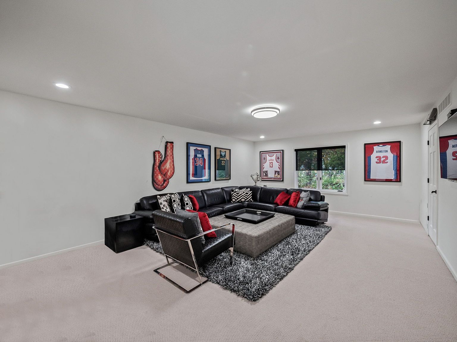 This is a well-decorated living room featuring a large, black leather sectional sofa with decorative pillows and a gray ottoman. The room is illuminated by recessed lighting and a ceiling light fixture. Sports memorabilia, including framed jerseys and boxing gloves, adorn the walls, adding a unique and personal touch to the space.