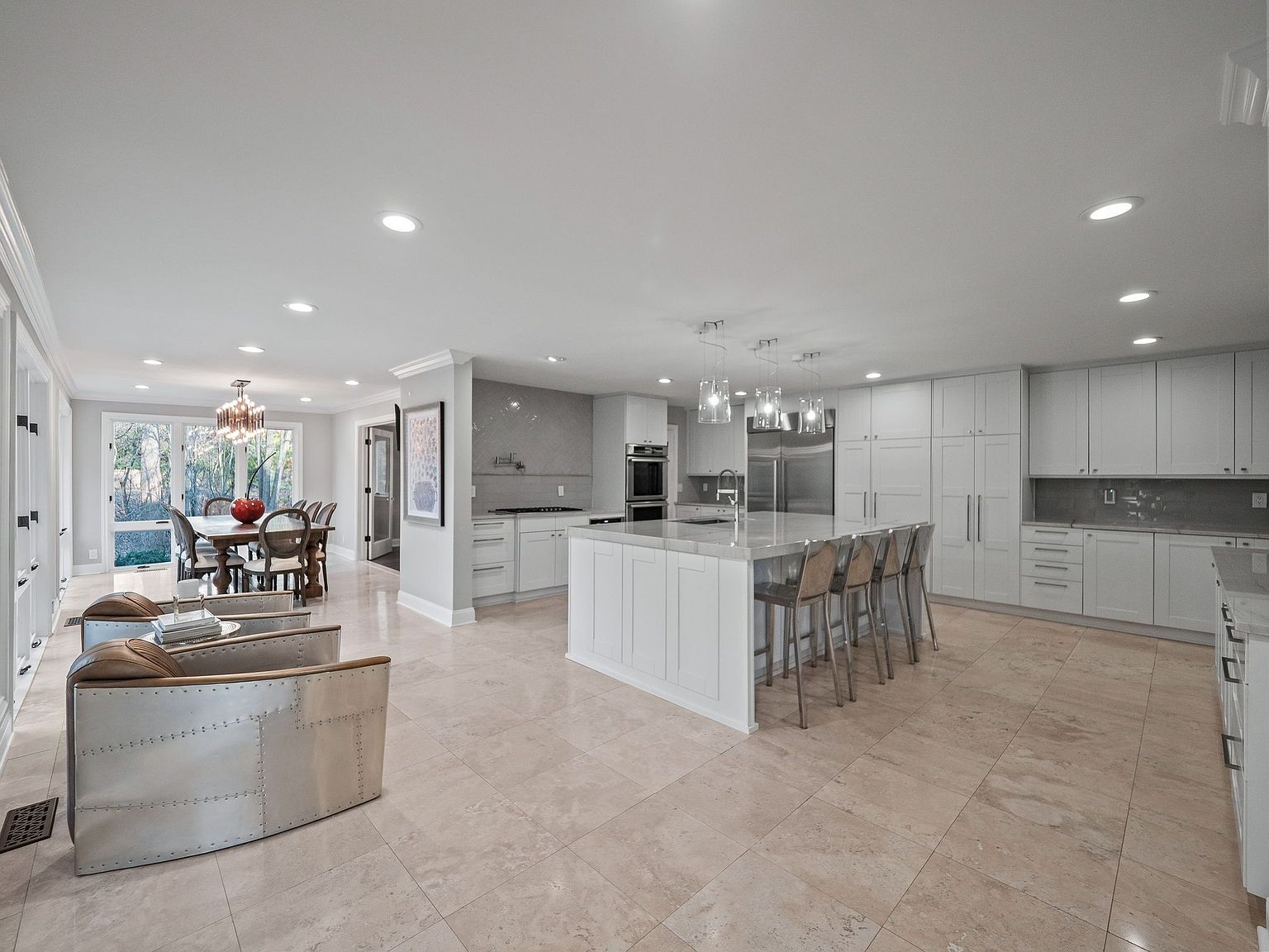 This is a spacious, open-concept kitchen and dining area featuring a large central island with bar seating, white cabinetry, and stainless steel appliances. The kitchen flows seamlessly into a dining area with a large table and chairs, and a seating area with unique metal-clad armchairs. The overall impression is one of modern elegance and functionality, perfect for both cooking and entertaining.
