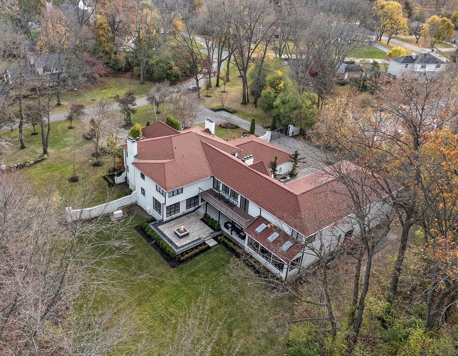 This aerial shot showcases a sprawling estate with a red-tiled roof and white exterior walls, surrounded by mature trees and lush landscaping. A modern patio with a fire pit adds an inviting outdoor space, while the overall design exudes luxury and privacy. The property is well-maintained and presents a grand appearance.