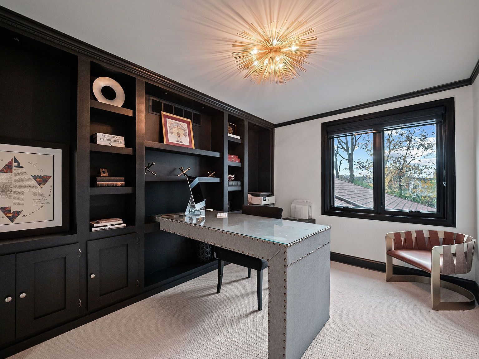 This is an interior shot of a sophisticated home office featuring a custom-built, dark-stained bookcase with open shelving and closed cabinets. A unique metal-clad desk with a glass top sits in front of the bookcase, complemented by a modern chair. A stylish light fixture illuminates the room, and a window provides natural light and a view of the outdoors.