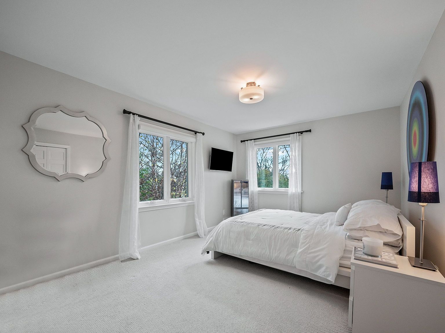 This is a bright and airy primary bedroom featuring a neutral color palette. The room has two windows with sheer white curtains, a bed with white linens, and a light-colored carpet. A decorative mirror hangs on the wall, and modern lamps flank the bed, creating a serene and inviting atmosphere.