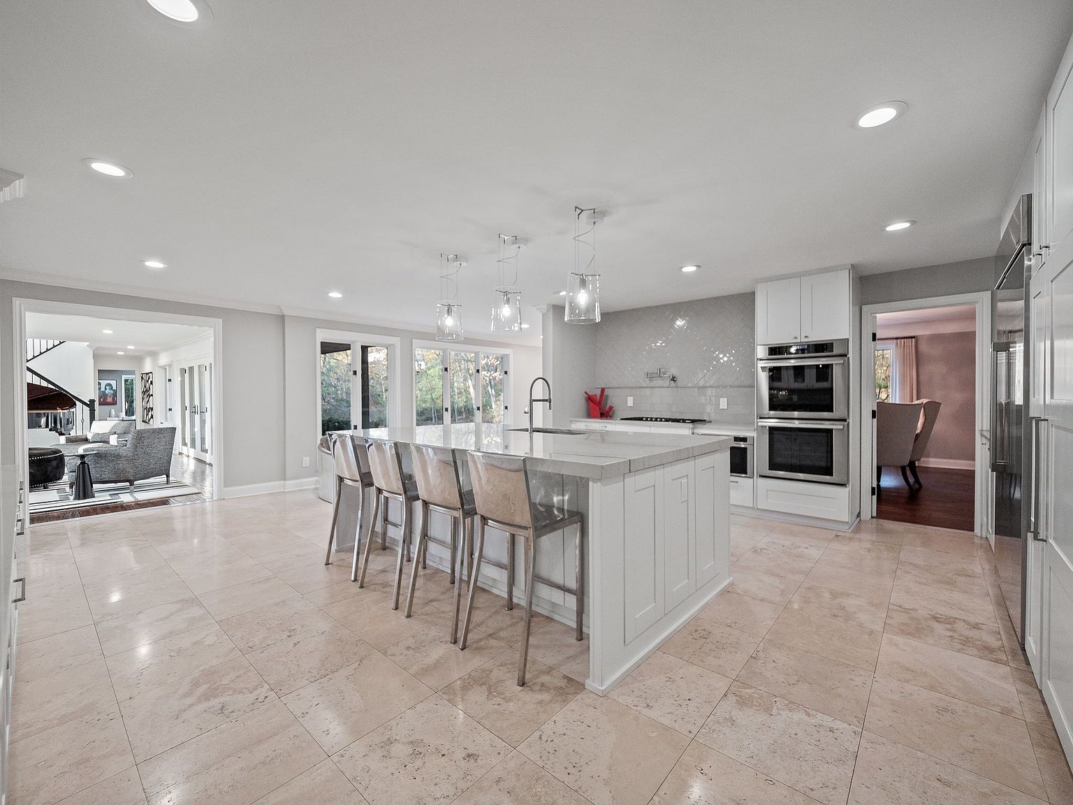 This is a bright and spacious kitchen featuring a large center island with seating, pendant lighting, and a light-colored countertop. The kitchen is equipped with stainless steel appliances, including double ovens, and has a neutral color palette with light tile flooring. The open layout connects to other living spaces, enhancing the home's flow and functionality.