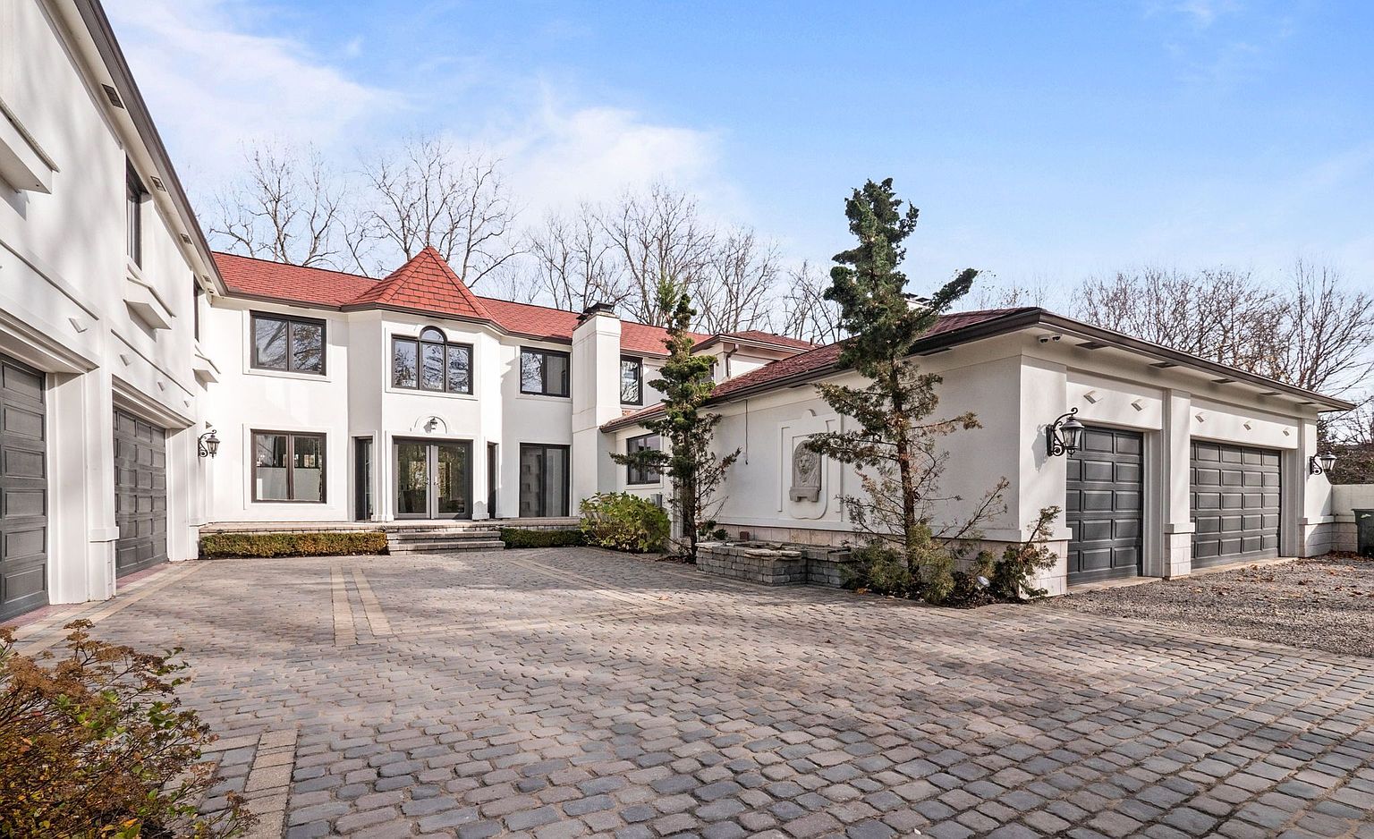 This is a front exterior view of a large, luxurious home with a red roof and white facade. The property features a spacious cobblestone driveway leading to multiple garage doors, complemented by manicured landscaping and mature trees. The architectural style is elegant and stately, creating a strong impression of upscale living.