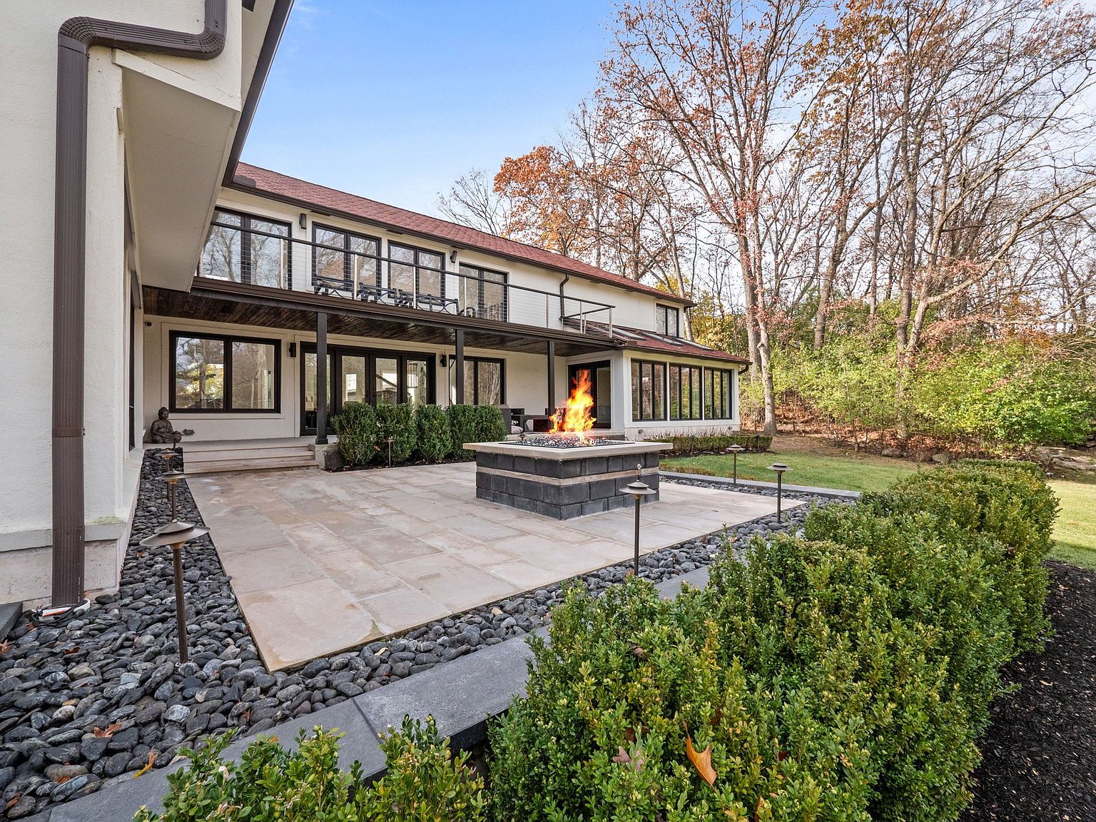 This image showcases a beautifully designed outdoor patio area featuring a modern fire pit as a focal point. The patio is constructed with large stone tiles, bordered by decorative rocks and lush greenery. The house, with its dark-framed windows and balcony, provides an elegant backdrop, creating an inviting and sophisticated outdoor living space.