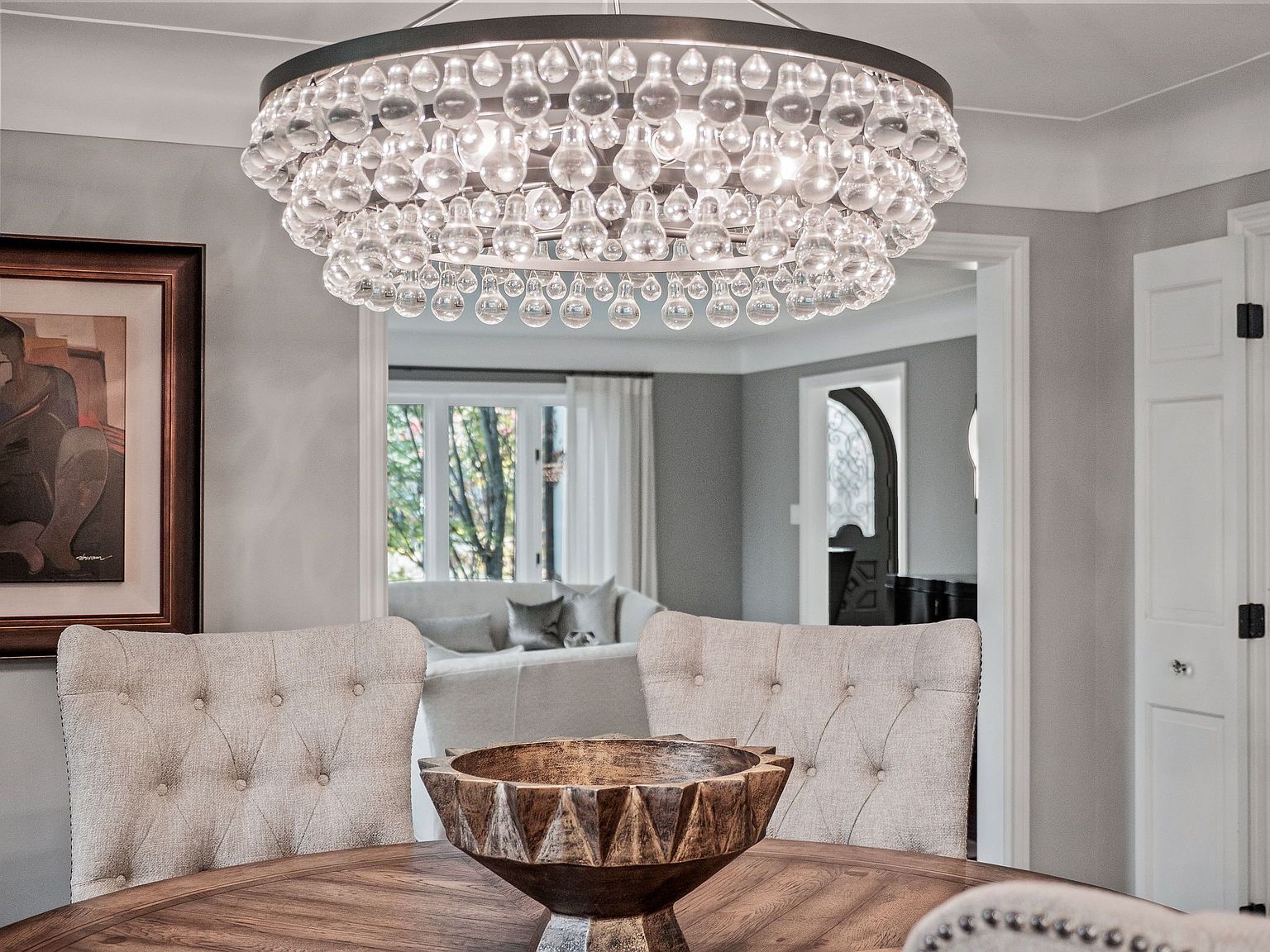 This is an interior shot of a dining room, showcasing a round wooden table with upholstered chairs. A large, ornate chandelier hangs above the table, providing ample lighting. The room is painted in neutral tones, and a glimpse into an adjacent living area reveals a comfortable sofa and natural light, creating an inviting and elegant atmosphere.