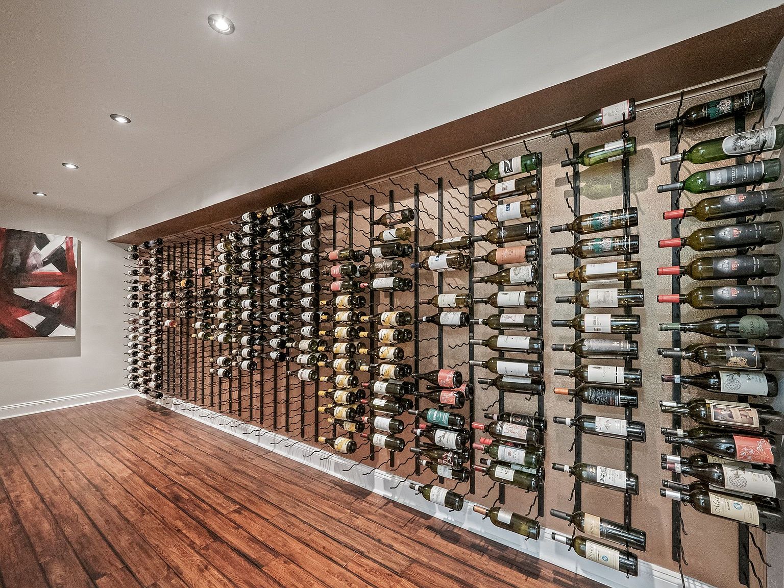 This is an interior shot of a well-stocked wine cellar. The walls are lined with racks holding numerous bottles of wine, creating an impressive display. The room features a hardwood floor and recessed lighting, contributing to a sophisticated and organized space perfect for wine enthusiasts.