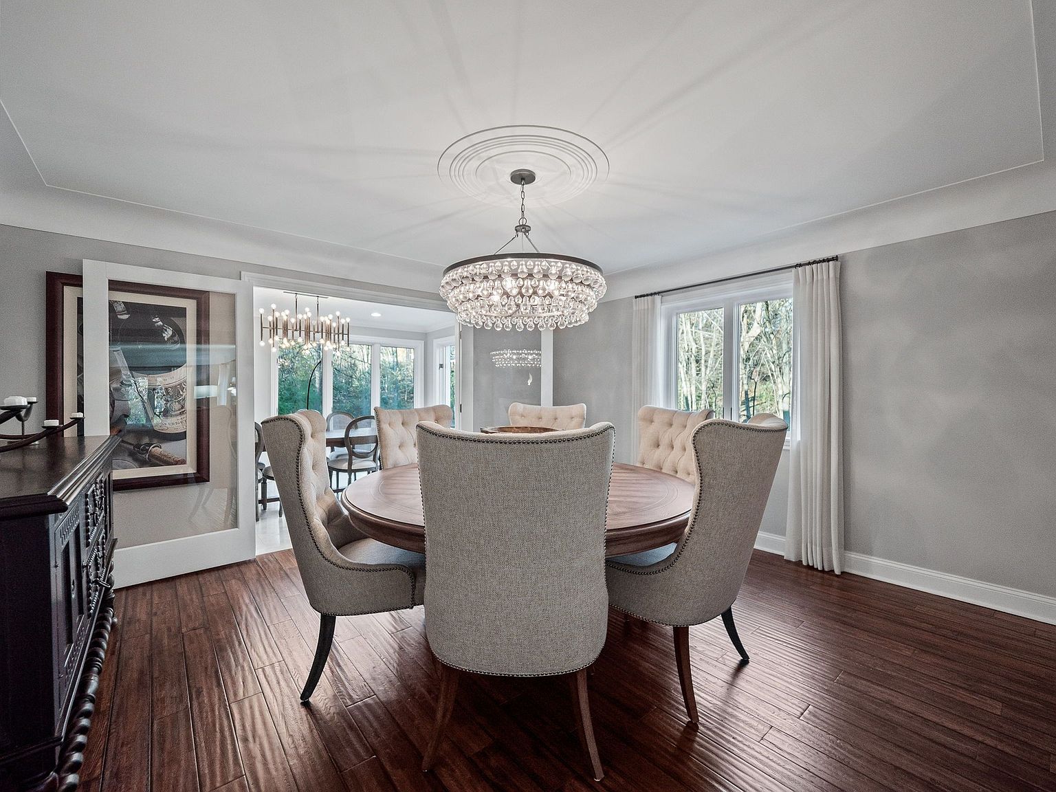 This is an interior shot of a dining room featuring a round wooden table surrounded by six upholstered chairs. A large, ornate chandelier hangs above the table, providing ample lighting. The room has hardwood floors and neutral-colored walls, creating a sophisticated and inviting atmosphere.