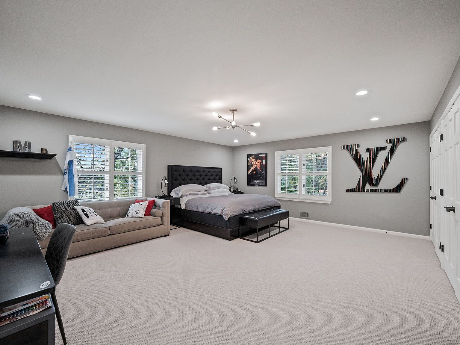 This is a spacious primary bedroom featuring a king-size bed with a tufted headboard, a comfortable sofa, and modern decor. The room is well-lit with natural light from the windows and recessed lighting, creating a bright and inviting atmosphere. A large LV logo art piece adds a unique touch to the room.
