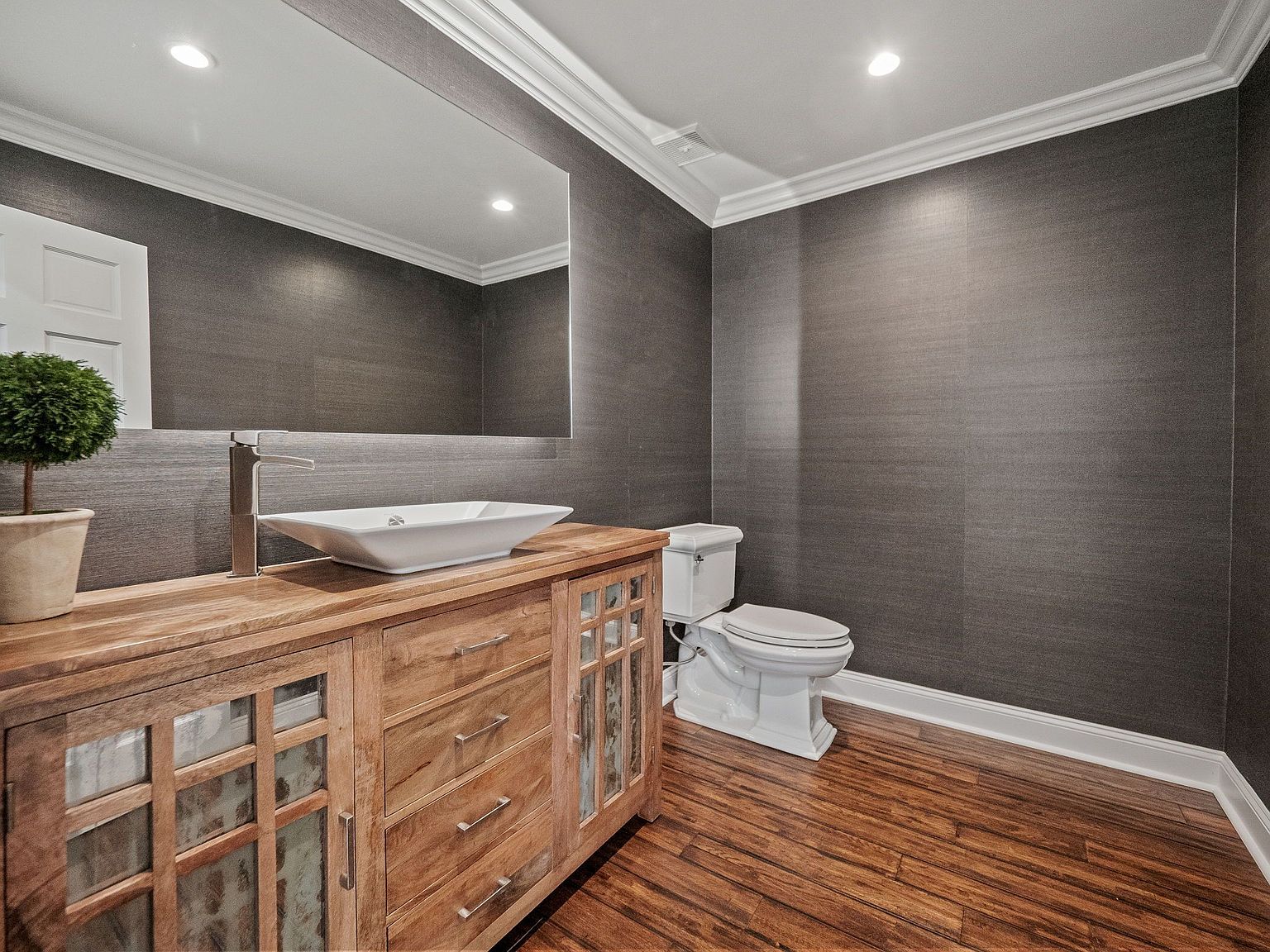This is a well-appointed guest bathroom featuring a wooden vanity with a vessel sink and modern faucet. The walls are covered in a textured gray wallpaper, complemented by white crown molding and baseboards. A toilet is visible to the right, and the flooring is a rich wood, creating a sophisticated and inviting space.