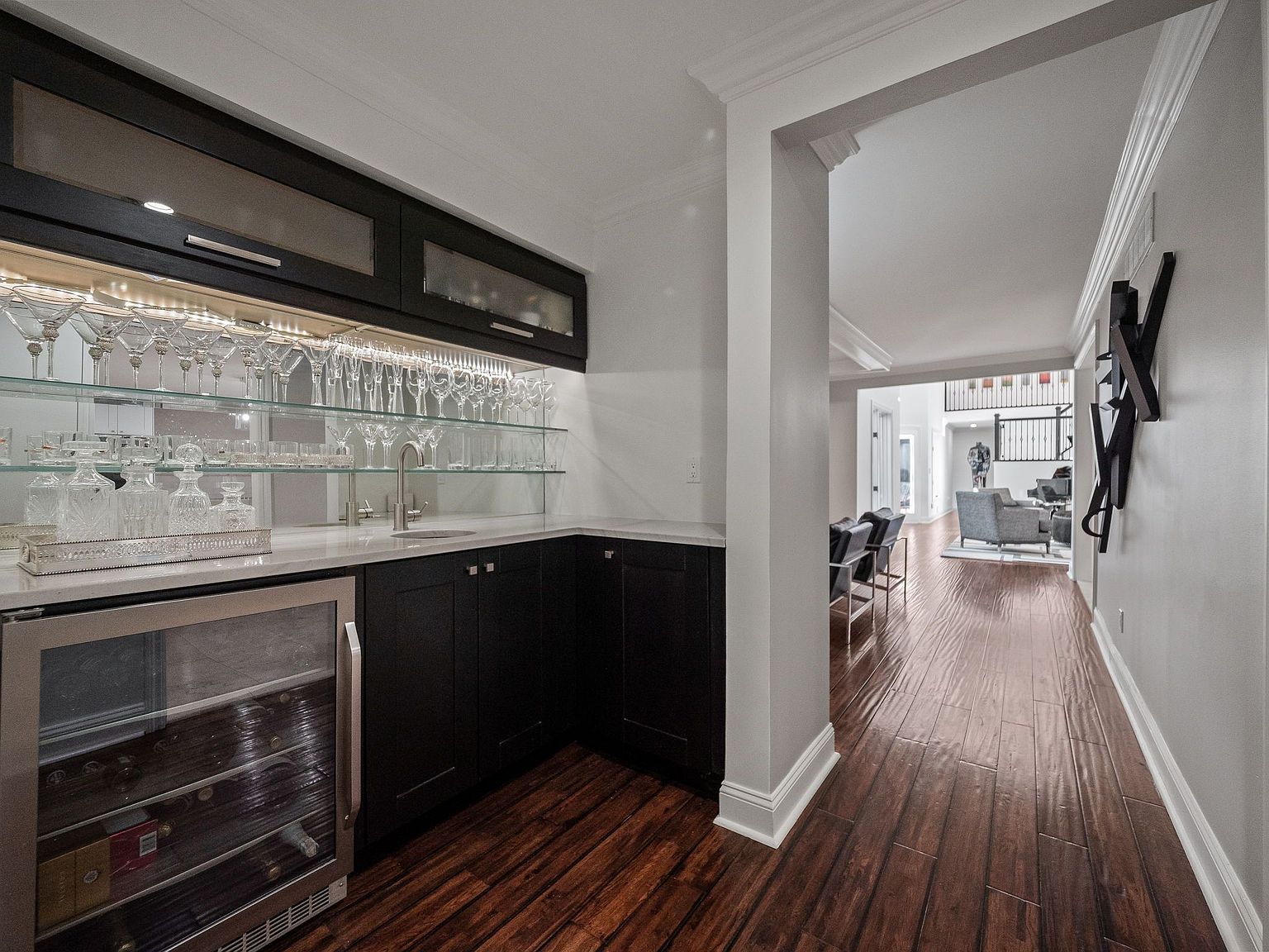 This image showcases a stylish wine cellar or bar area featuring dark cabinetry, glass shelving displaying various glassware, and a built-in wine refrigerator. The space is well-lit, highlighting the collection of bottles and glasses, and the dark wood flooring adds a touch of elegance. The view extends into a hallway and living area, suggesting a seamless flow within the home.