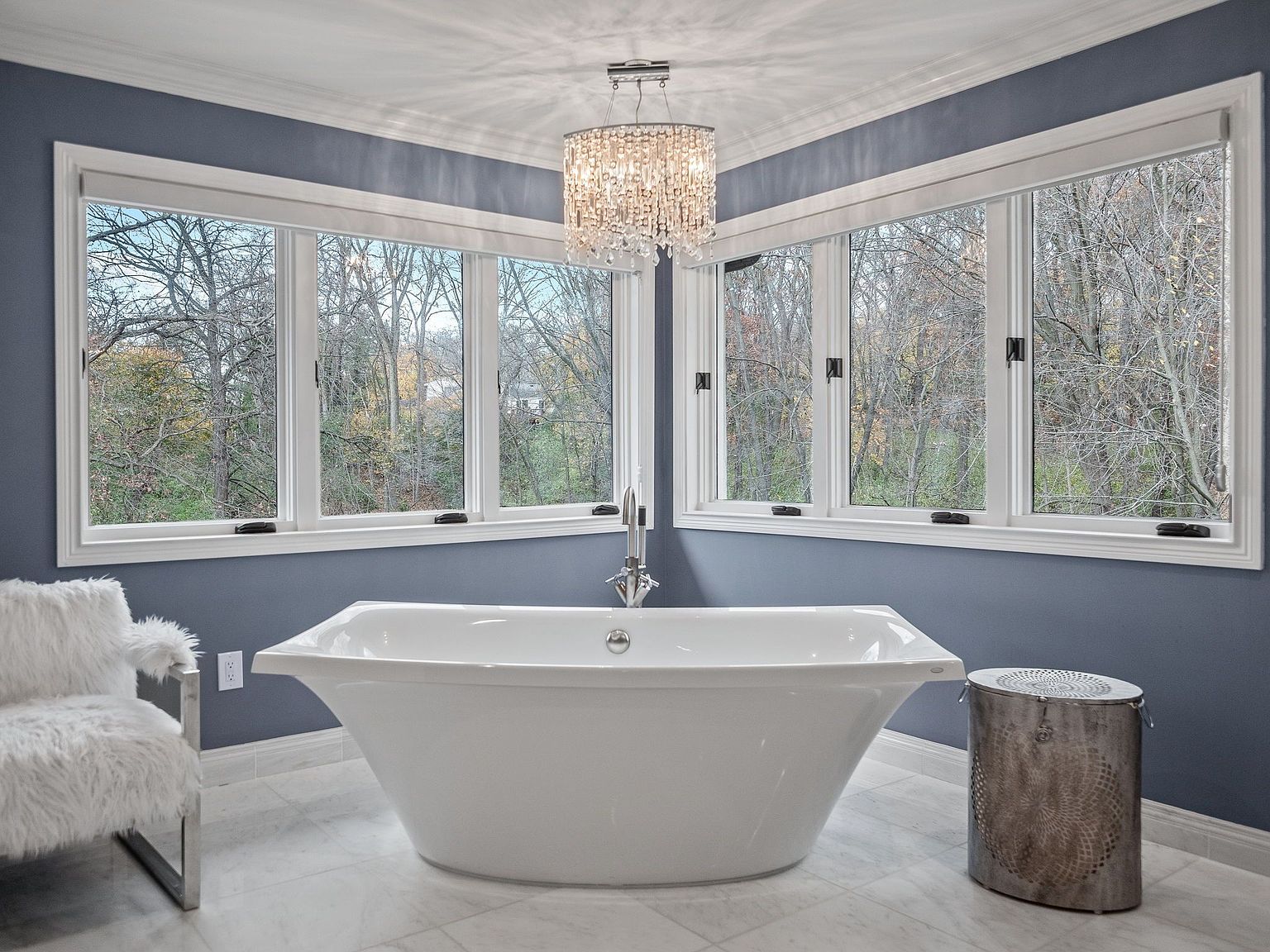 This is a luxurious primary bathroom featuring a freestanding white bathtub positioned in front of a corner window offering a view of trees. A crystal chandelier hangs above the tub, adding a touch of elegance. The walls are painted a calming blue, and the floor is tiled with a light-colored stone, creating a serene and spa-like atmosphere.