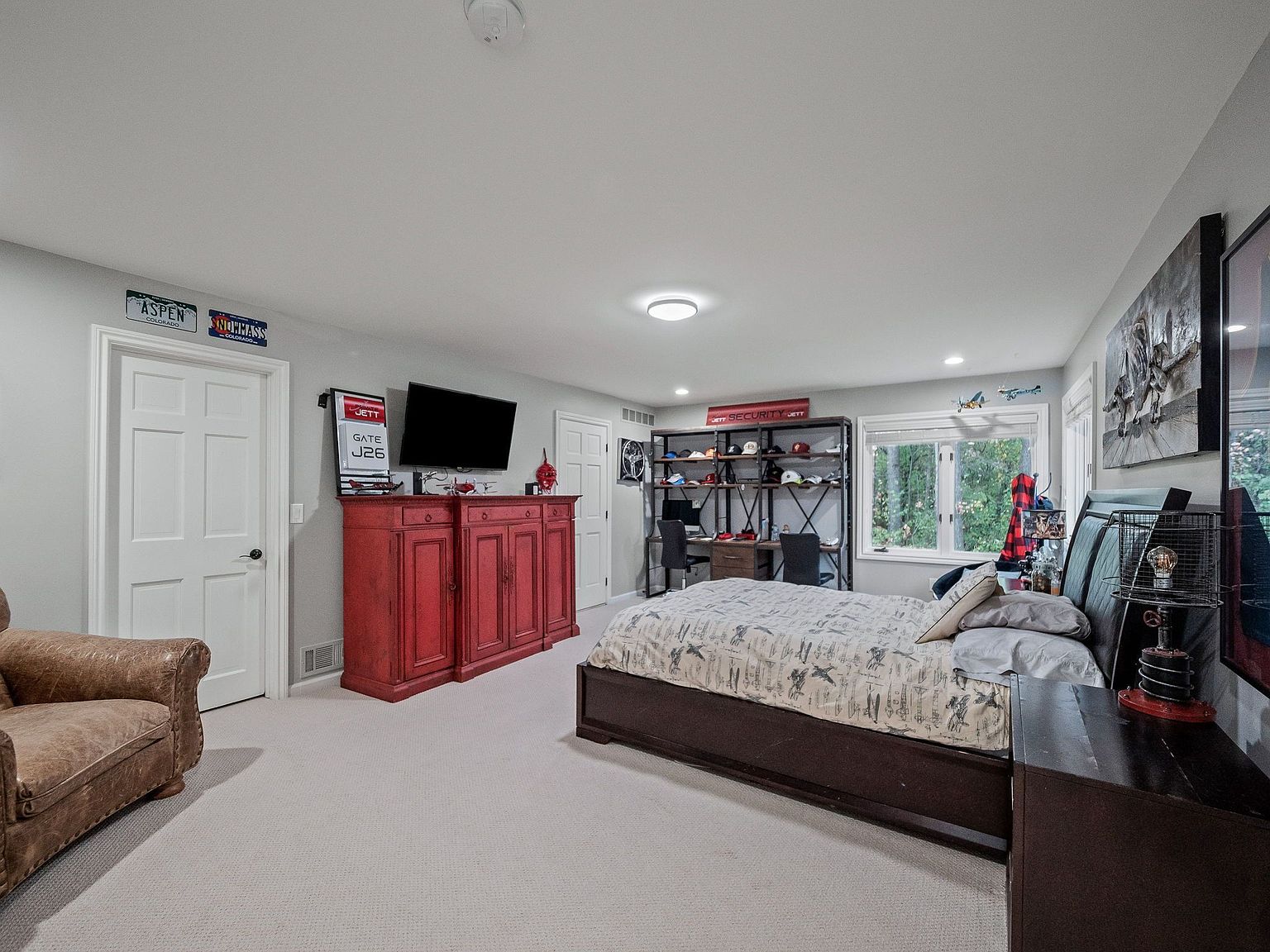 This is a well-appointed bedroom featuring a dark wood bed frame with a patterned comforter, a matching nightstand, and a red cabinet under a wall-mounted TV. A large window provides natural light, and a bookshelf with hats and decorative items adds character. The room has a cozy and inviting atmosphere, suitable for a guest or child.