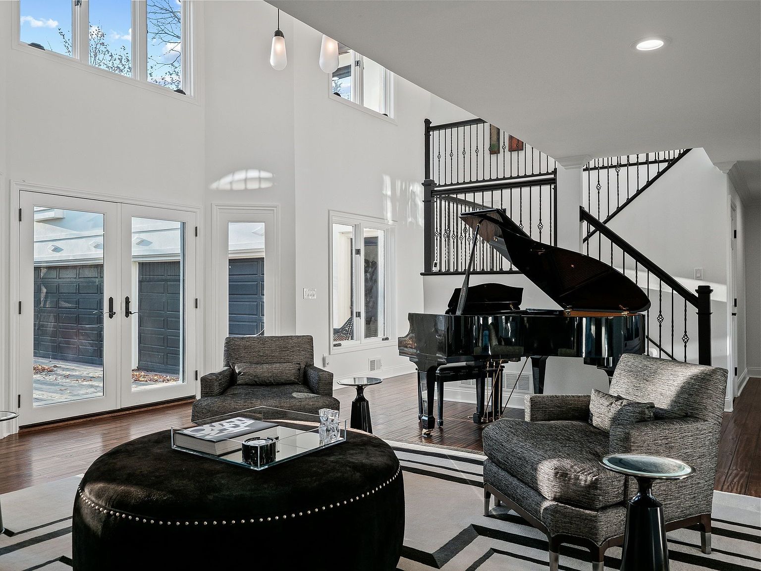 This is an interior shot of a modern living room featuring a grand piano, two armchairs, and a large ottoman. The room has high ceilings, large windows, and a staircase with black metal railings. The overall impression is elegant and spacious, perfect for entertaining.