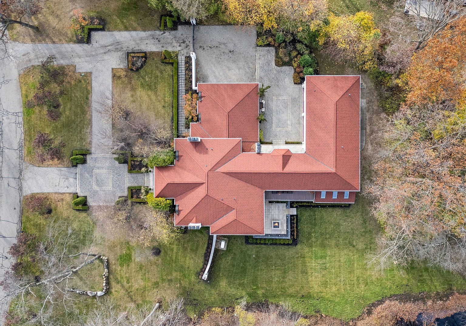 This aerial view showcases a large, luxurious home with a red tile roof, surrounded by meticulously landscaped grounds and mature trees. The property features multiple driveways, manicured lawns, and a patio area with a fire pit. The overall impression is one of grandeur and privacy, highlighting the expansive estate.