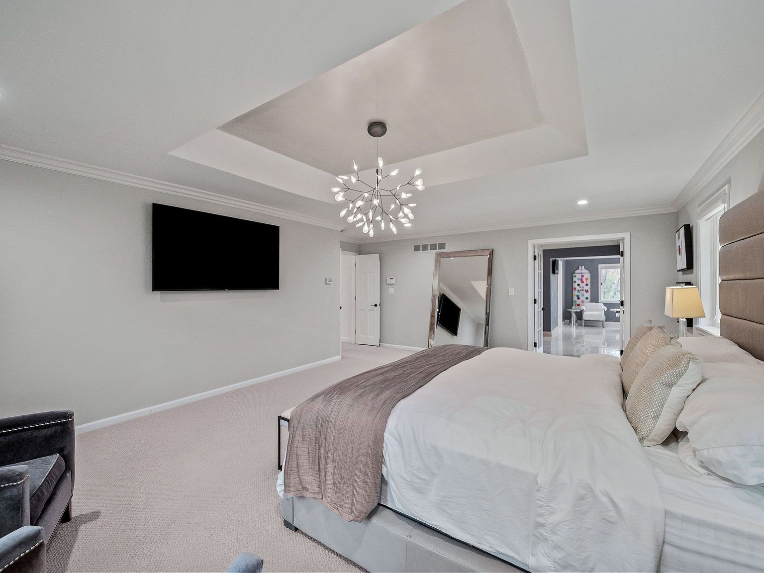 This is a primary bedroom featuring a large bed with a padded headboard, white linens, and a taupe throw. The room has neutral-colored walls, carpeted floors, and a modern chandelier. A large television is mounted on the wall, and a full-length mirror stands near the doorway, reflecting light and adding depth to the space.