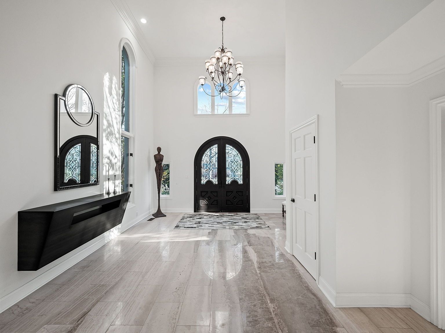 This is an interior shot of a grand hallway featuring high ceilings, a decorative chandelier, and a dark wood front door with ornate glass detailing. A modern floating console table with a mirror above it sits against one wall, while a sculpture stands near a tall arched window. The flooring is light-colored tile, and the overall impression is one of spaciousness and luxury.