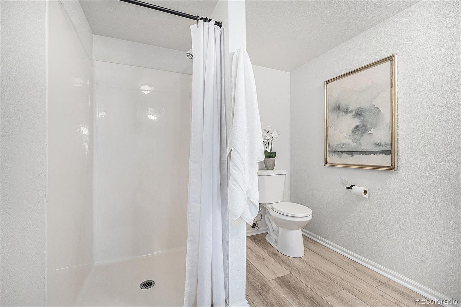 This is a bright and clean guest bathroom featuring a white shower stall with a black shower rod and white curtain, a white toilet with a small potted orchid on top, and a framed abstract painting on the wall. The flooring is light wood-look laminate, and the walls are a light neutral color, creating a fresh and inviting space.