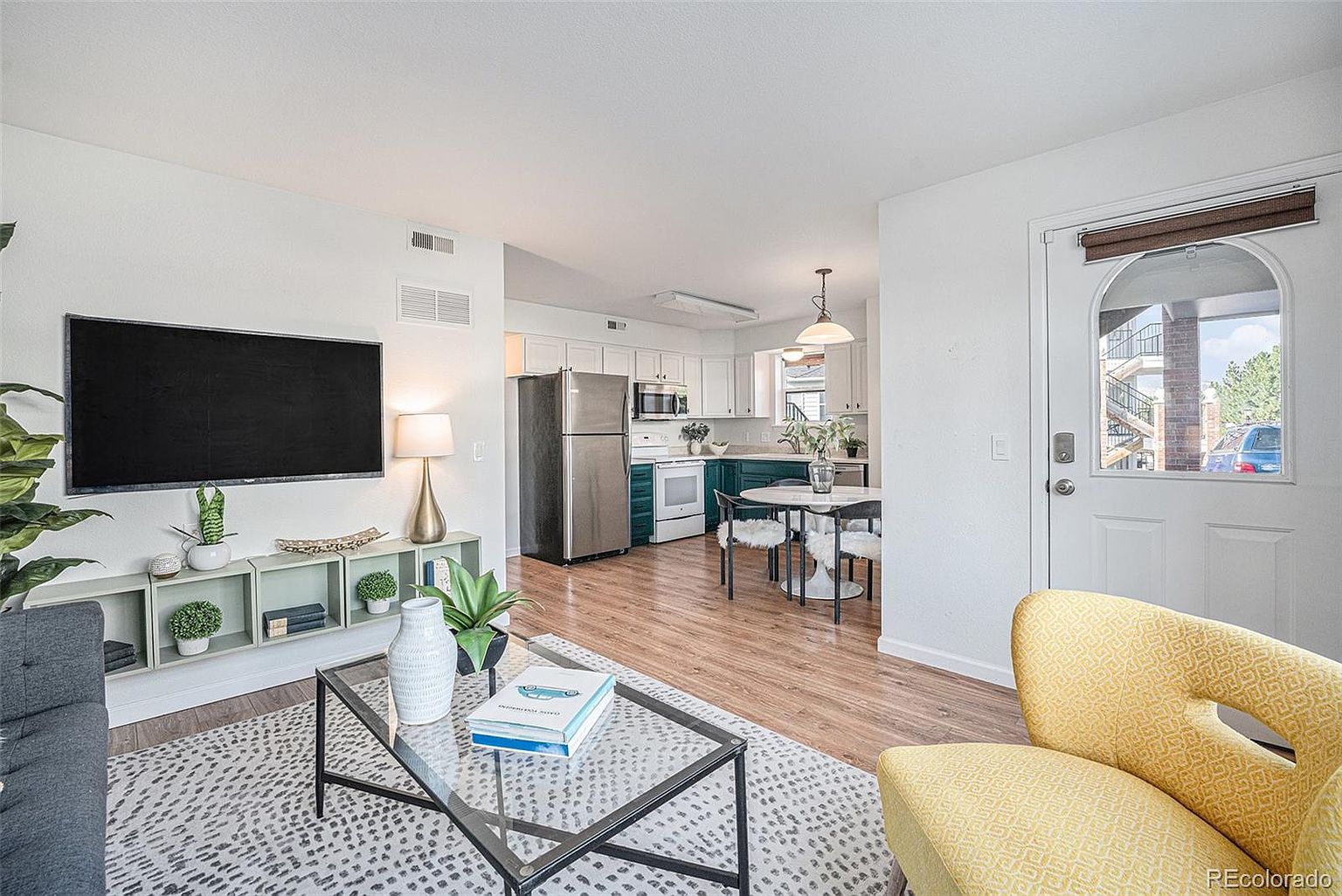 This is an interior shot of a living room that flows into a kitchen and dining area. The living room features a television mounted on the wall above a low shelving unit, a glass coffee table on a patterned rug, and a yellow patterned armchair. The kitchen has white cabinets and stainless steel appliances, and the dining area includes a round table with chairs. The overall impression is a bright and open living space.