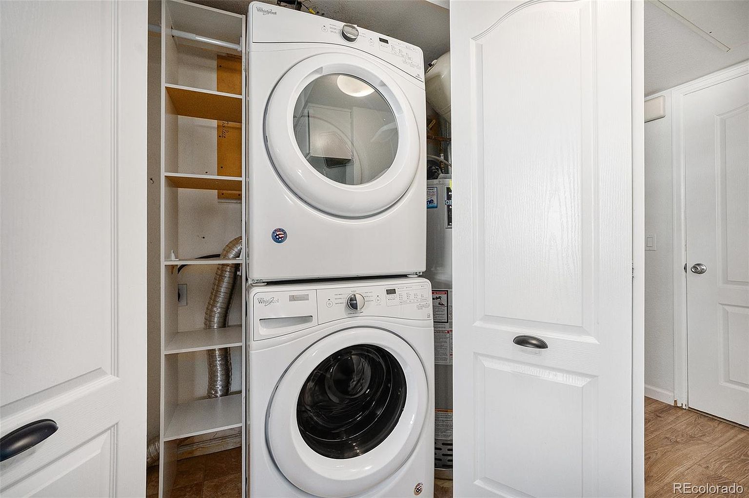 This image showcases a compact laundry area featuring a stacked Whirlpool washer and dryer set. Adjacent to the appliances is a built-in shelving unit, providing storage space. A closed white door is visible on the right, suggesting a clean and organized space.