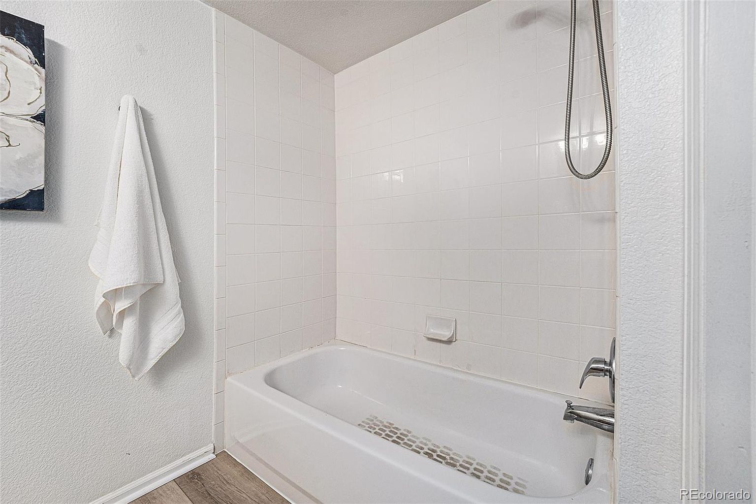 This is a clean and bright bathroom featuring a white bathtub with tiled walls and a shower head. A white towel hangs on the wall next to an abstract art piece. The floor is a light wood-look laminate, and the overall impression is fresh and well-maintained.