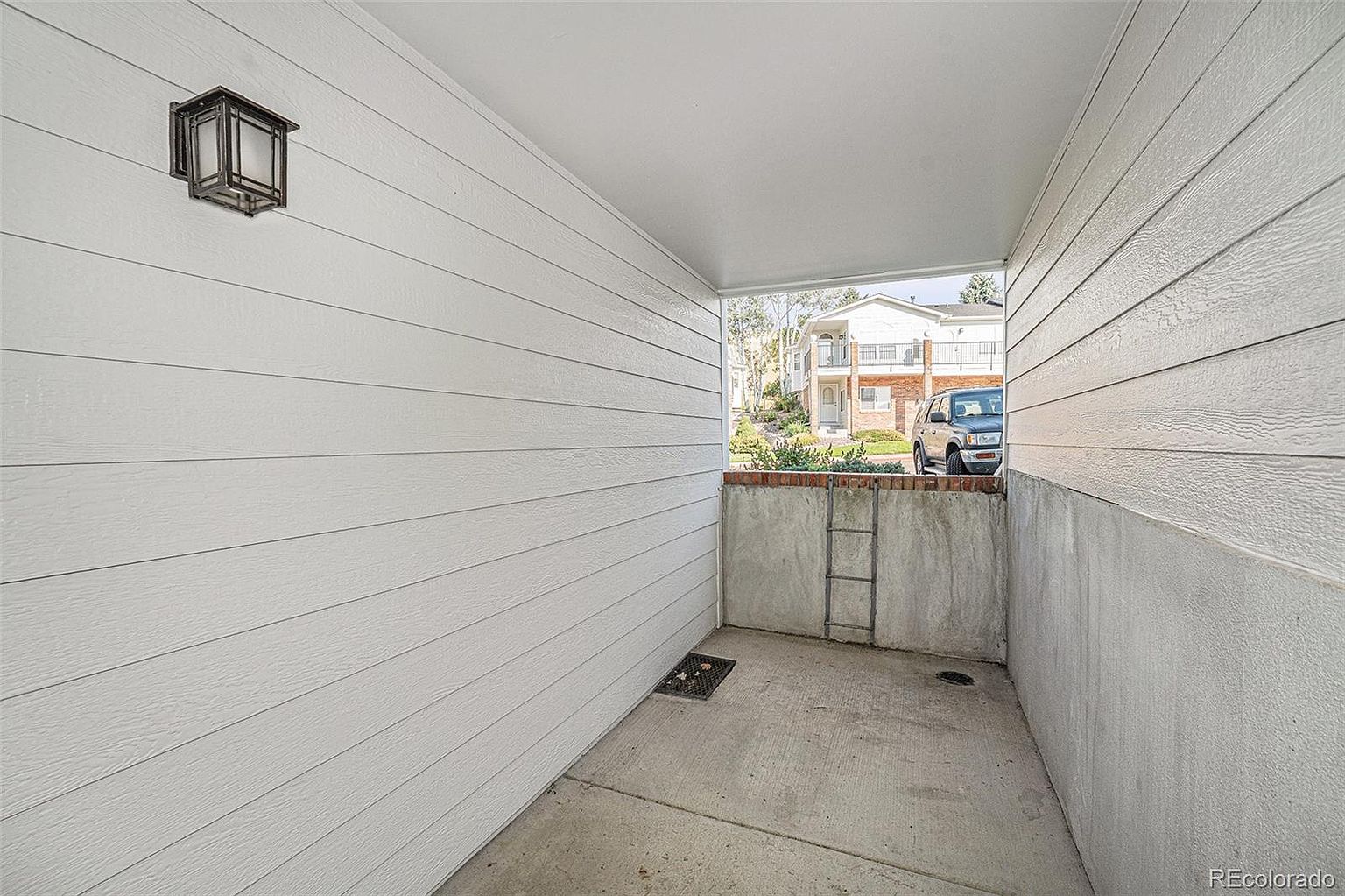 This image showcases a covered entryway with white horizontal siding and a concrete floor. A modern, dark-colored lantern-style light fixture is mounted on the wall. The entryway leads to an exterior view of a building and a parked vehicle, suggesting a welcoming and functional space.