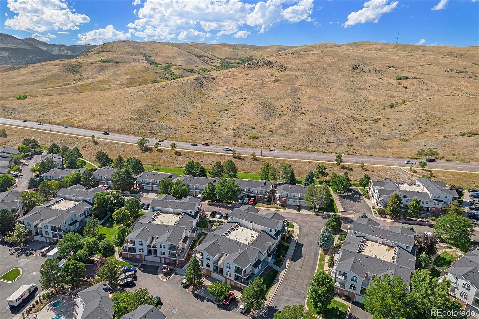 This aerial view showcases a well-maintained residential complex with multiple buildings featuring gray roofs and light-colored facades. Lush green trees surround the buildings, adding to the aesthetic appeal. A road with light traffic runs adjacent to the complex, with a backdrop of rolling hills and a clear blue sky with scattered clouds, creating a serene and desirable living environment.